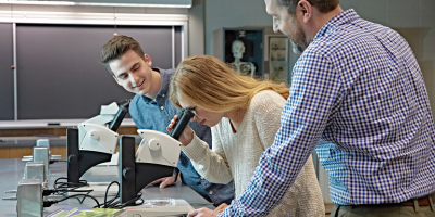 A teacher instructs two students using microscopes in a science classroom. A skeleton model is visible in the background.