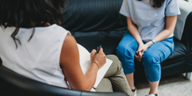 Two people in casual clothing sit on a dark sofa and on the floor while one person writes in a notebook with a pen, suggesting a calm conversation or counseling moment.