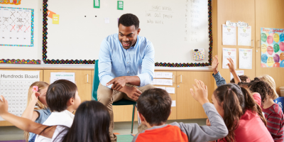A male teacher with a blue button down sits in front of a white board. A group of children with their hands raised sit in front of him on the floor.