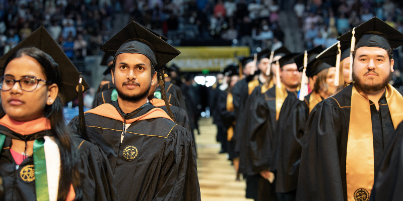 Two lines of students in graduate and undergraduate regalia stare at the camera. In the background, stadium seating and a crowd sit watching the graduation ceremony.