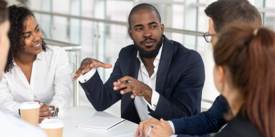 A group of people in business attire are seated around a table, engaged in a discussion. One person is gesturing with their hands as they speak, while others listen attentively. Coffee cups and a notebook are visible on the table.