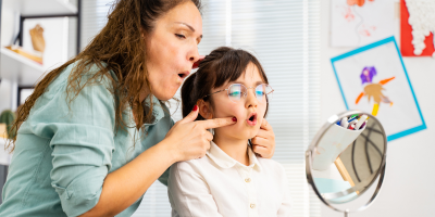 A speech therapist guides a child in front of a mirror, demonstrating mouth exercises.